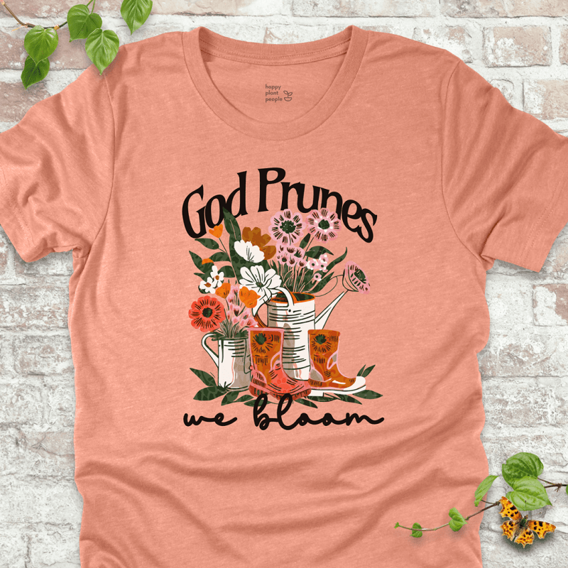 Peach "We Bloom" T-shirt featuring illustrated rain boots, watering cans, and blooming flowers with the text "God Prunes, We Bloom."