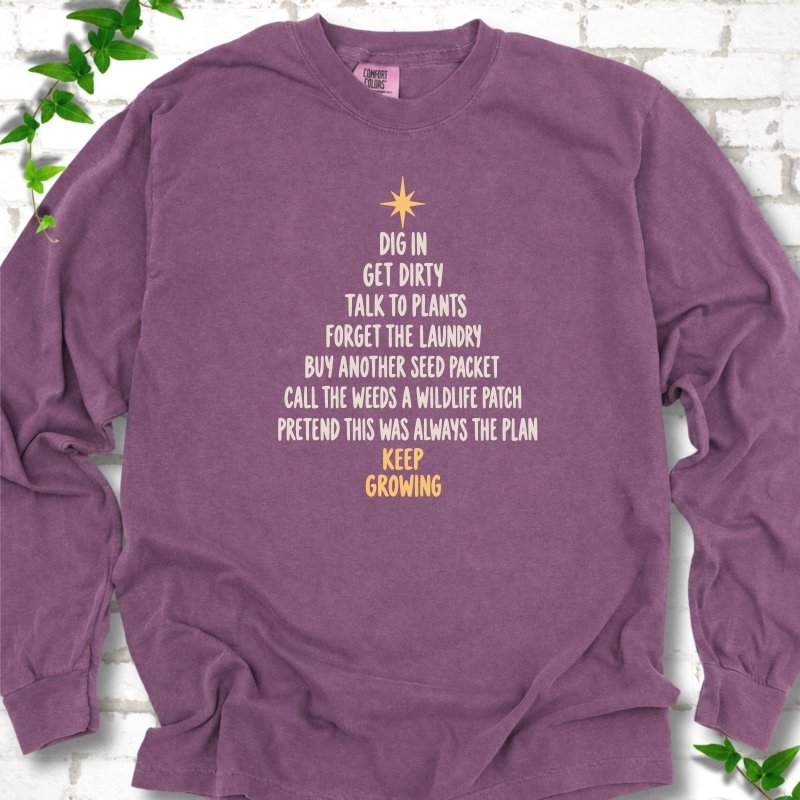 Berry purple long sleeve T-shirt with a Christmas tree made of cream hand-lettered gardening phrases topped by a gold star, flat lay on white brick with ivy
