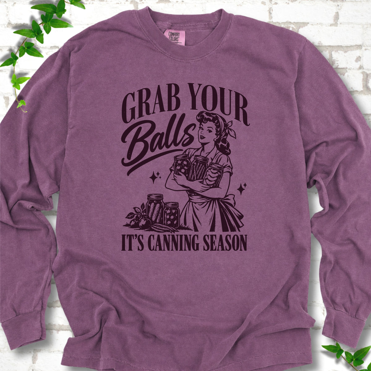 Berry long sleeve T-shirt flat lay with a dark plum retro illustration of a 1950s-style woman in a headscarf and apron hugging three filled canning jars, more jars and vegetables at her feet, with "Grab your" above, "Balls" in script and "It's canning season" below, on white brick with ivy.