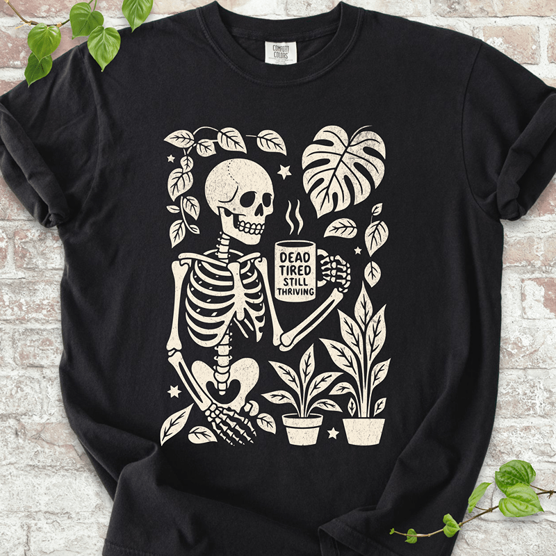 Dead Tired T-shirt on Comfort Colors® Black – shows a skeleton graphic sipping coffee among plants and stars with Monstera leaves in a high-contrast print