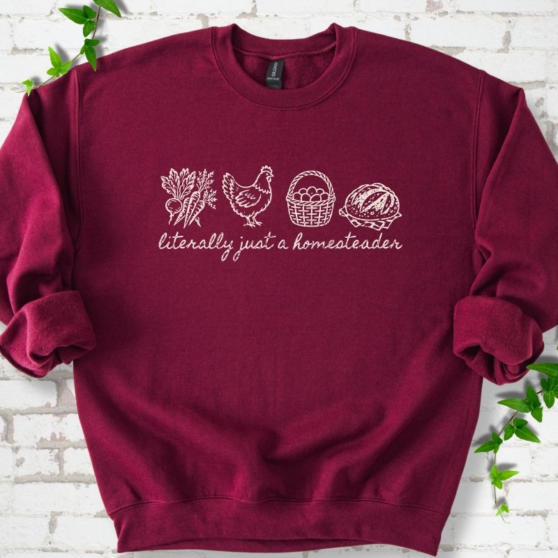 Maroon sweatshirt flat lay with white line-art icons in a row of carrots and radishes, a hen, a basket of eggs and a loaf of bread above the script words "literally just a homesteader", on white brick with ivy sprigs.