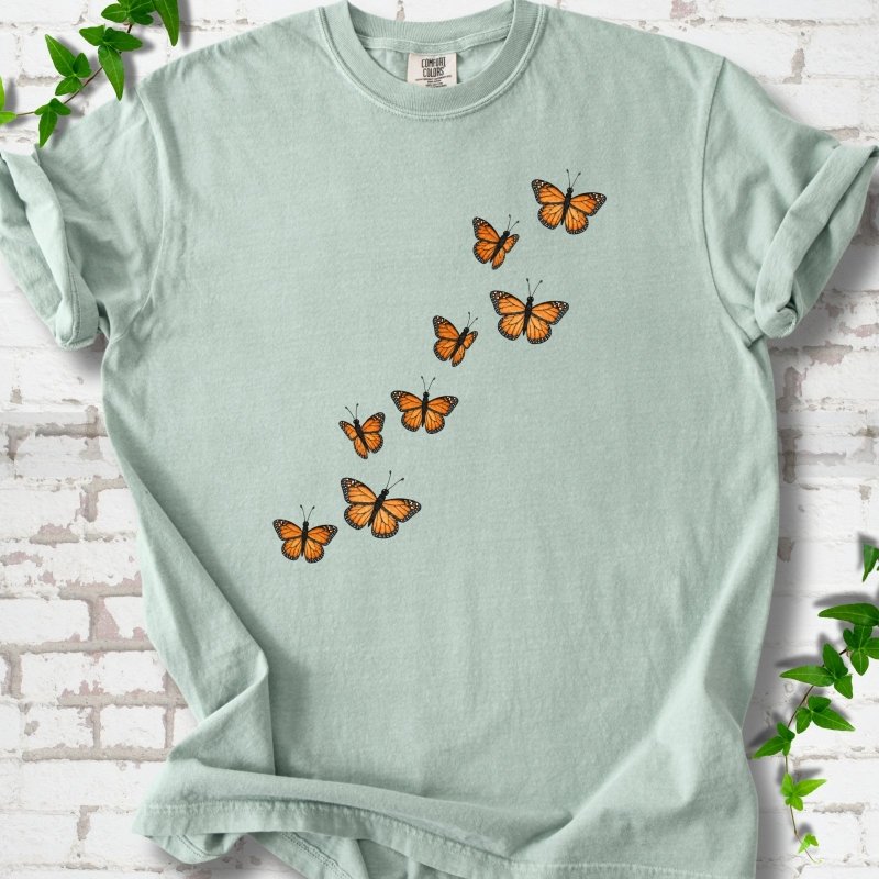 Bay light green short-sleeve T-shirt with eight orange monarch butterflies flying in a rising diagonal trail across the chest, flat lay on white brick with ivy