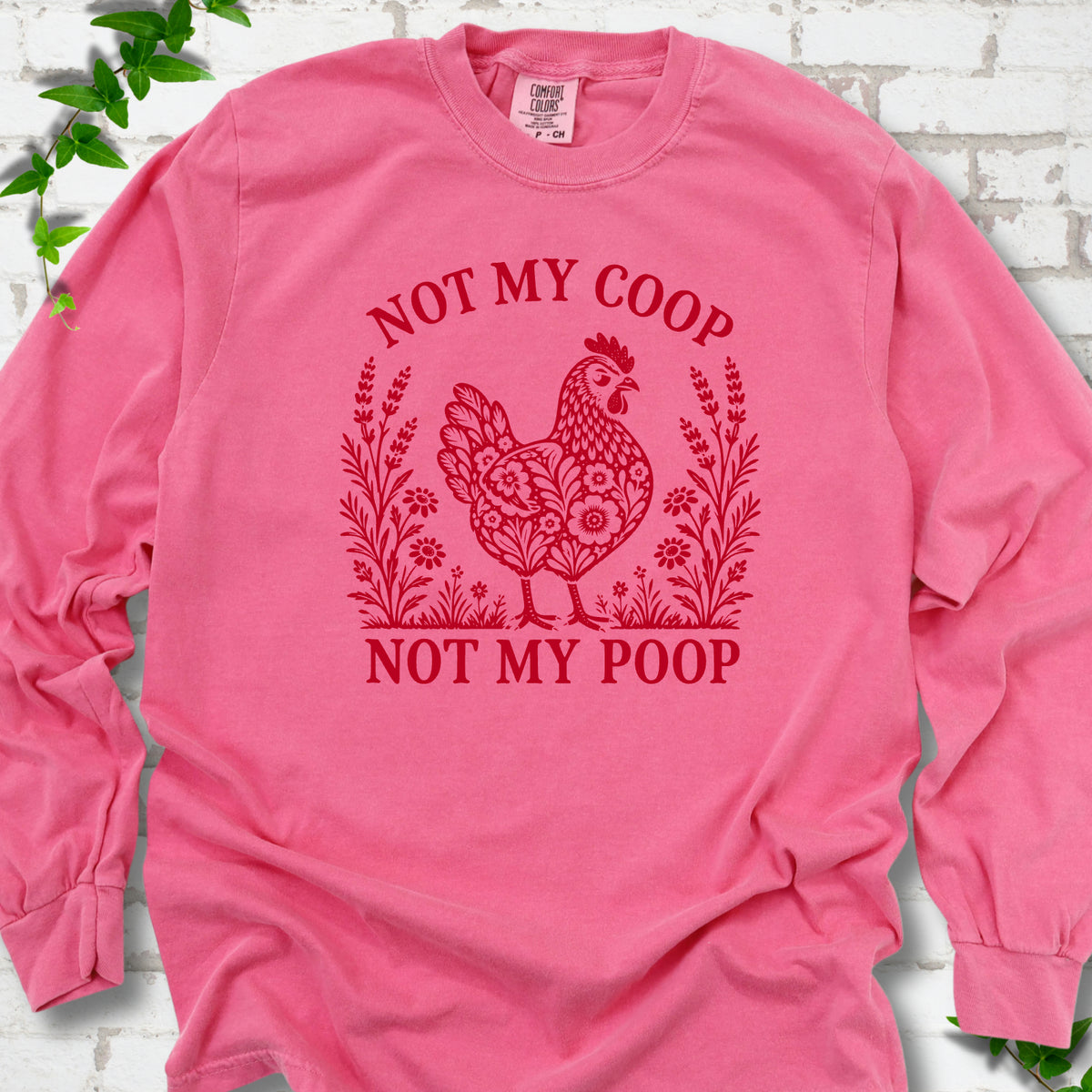 Crunchberry long sleeve T-shirt flat lay with a bright red folk-art hen surrounded by wildflowers and lavender, "Not my coop" arched above and "Not my poop" below, on white brick with ivy.