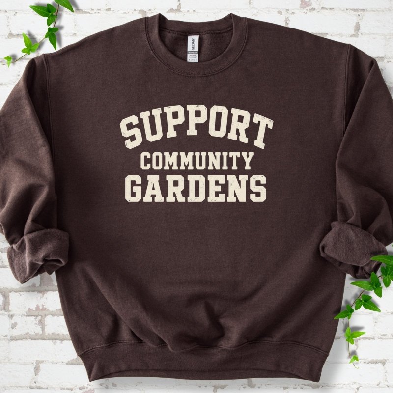 Dark chocolate brown crewneck sweatshirt with Support Community Gardens in cream collegiate block letters across the chest, flat lay on white brick with ivy