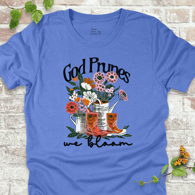 Blue "We Bloom" T-shirt featuring illustrated rain boots, watering cans, and blooming flowers with the text "God Prunes, We Bloom."