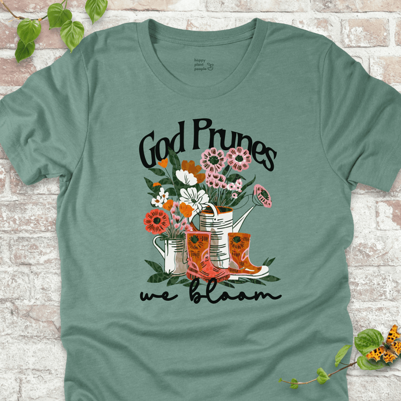 Dusty Blue "We Bloom" T-shirt featuring illustrated rain boots, watering cans, and blooming flowers with the text "God Prunes, We Bloom."