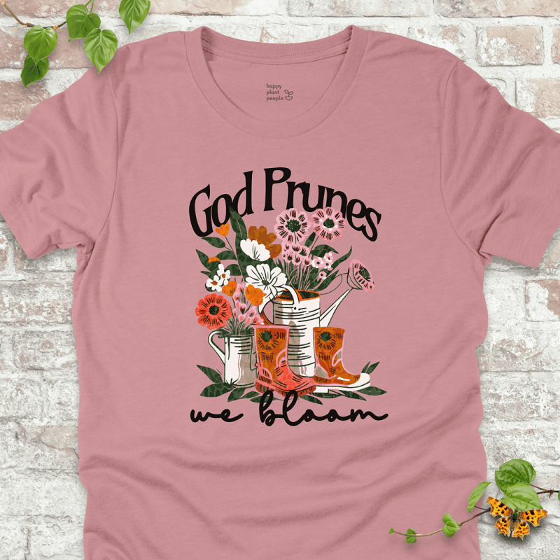 Pink "We Bloom" T-shirt featuring illustrated rain boots, watering cans, and blooming flowers with the text "God Prunes, We Bloom."