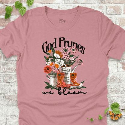 Pink "We Bloom" T-shirt featuring illustrated rain boots, watering cans, and blooming flowers with the text "God Prunes, We Bloom."
