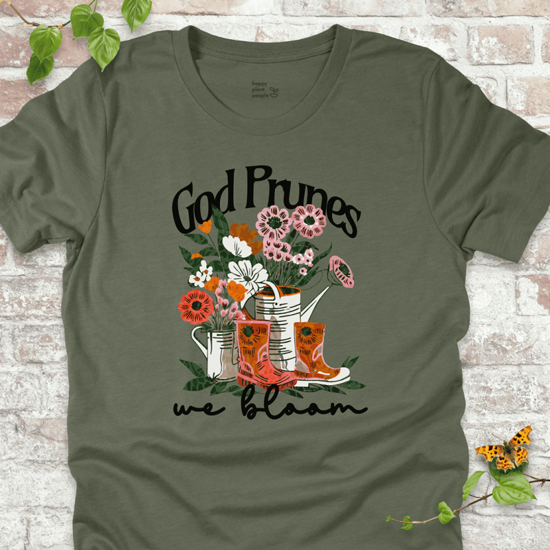 Green "We Bloom" T-shirt featuring illustrated rain boots, watering cans, and blooming flowers with the text "God Prunes, We Bloom."