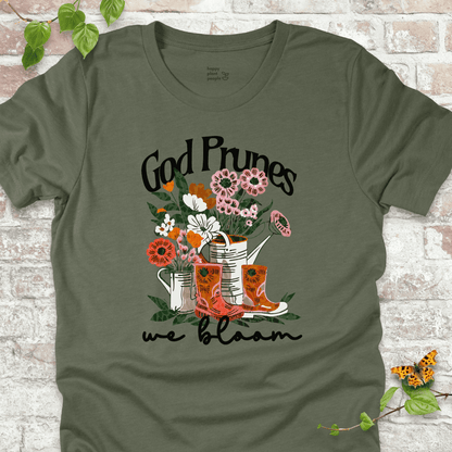 Green "We Bloom" T-shirt featuring illustrated rain boots, watering cans, and blooming flowers with the text "God Prunes, We Bloom."