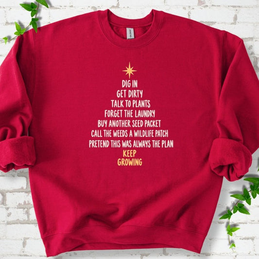 Cardinal red crewneck sweatshirt with a Christmas tree made of cream hand-lettered gardening phrases topped by a gold star, flat lay on white brick with ivy