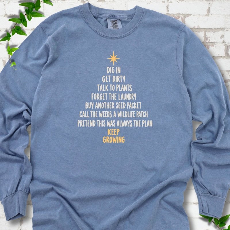 Blue Jean light blue long sleeve T-shirt with a Christmas tree made of cream hand-lettered gardening phrases topped by a gold star, flat lay on white brick with ivy