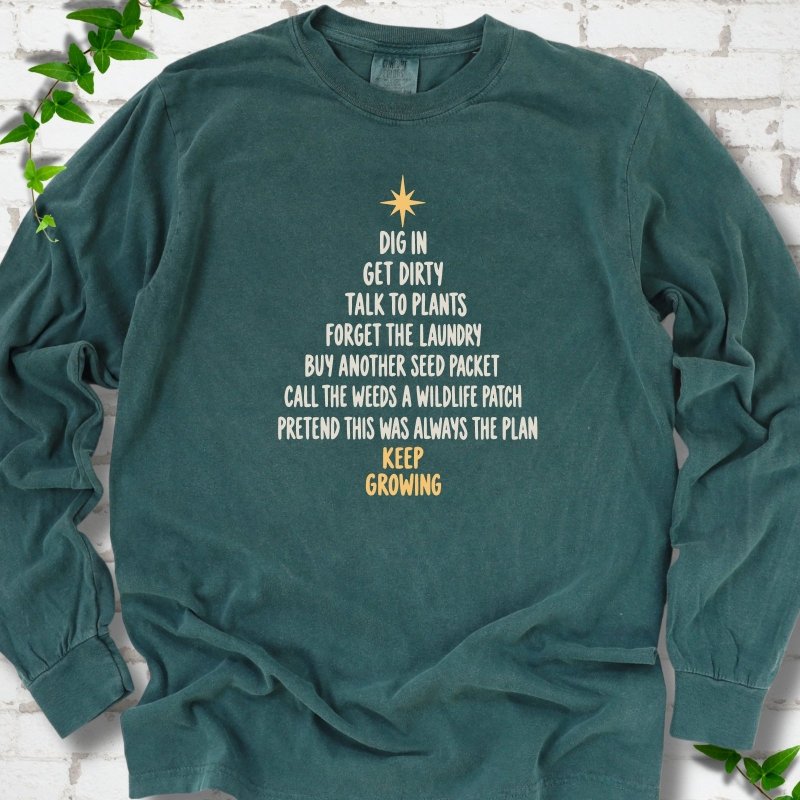 Blue Spruce green long sleeve T-shirt with a Christmas tree made of cream hand-lettered gardening phrases topped by a gold star, flat lay on white brick with ivy