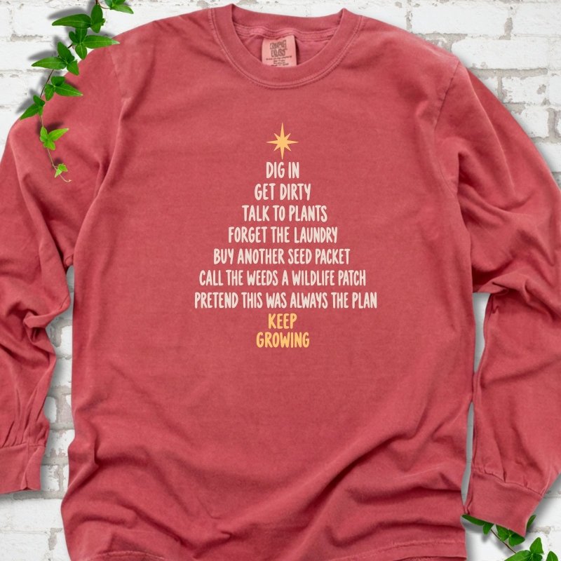 Crimson red long sleeve T-shirt with a Christmas tree made of cream hand-lettered gardening phrases topped by a gold star, flat lay on white brick with ivy
