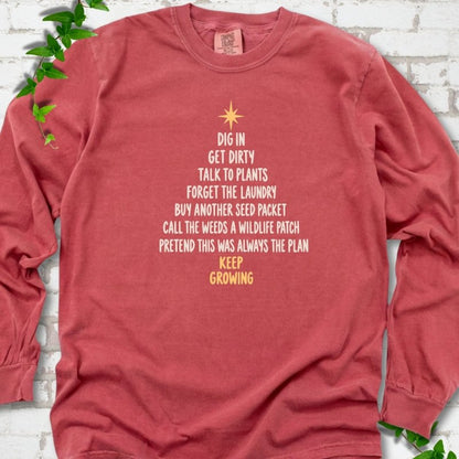 Crimson red long sleeve T-shirt with a Christmas tree made of cream hand-lettered gardening phrases topped by a gold star, flat lay on white brick with ivy