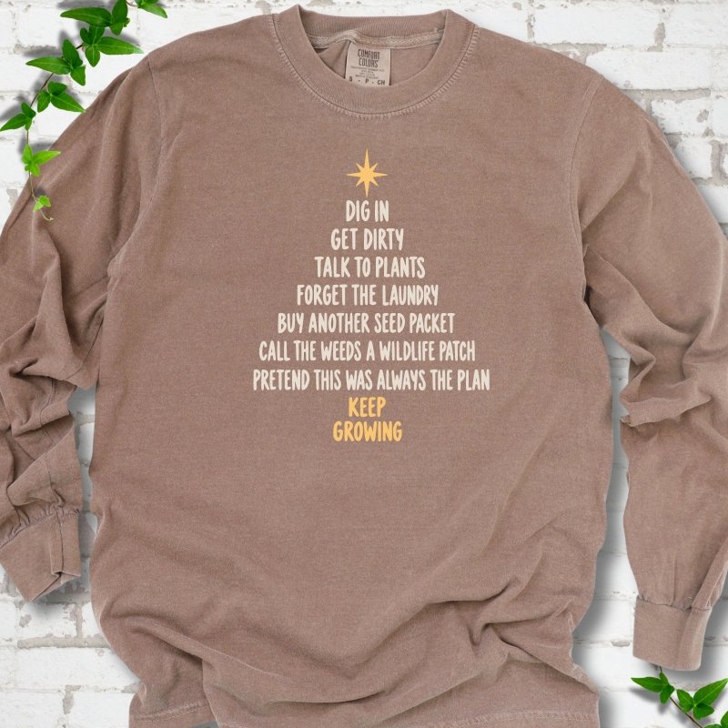 Espresso brown long sleeve T-shirt with a Christmas tree made of cream hand-lettered gardening phrases topped by a gold star, flat lay on white brick with ivy