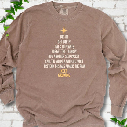 Espresso brown long sleeve T-shirt with a Christmas tree made of cream hand-lettered gardening phrases topped by a gold star, flat lay on white brick with ivy
