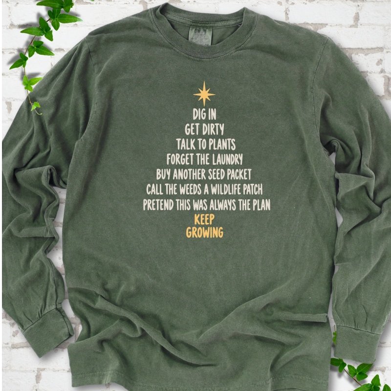 Hemp olive green long sleeve T-shirt with a Christmas tree made of cream hand-lettered gardening phrases topped by a gold star, flat lay on white brick with ivy