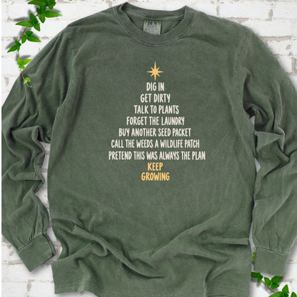 Hemp olive green long sleeve T-shirt with a Christmas tree made of cream hand-lettered gardening phrases topped by a gold star, flat lay on white brick with ivy