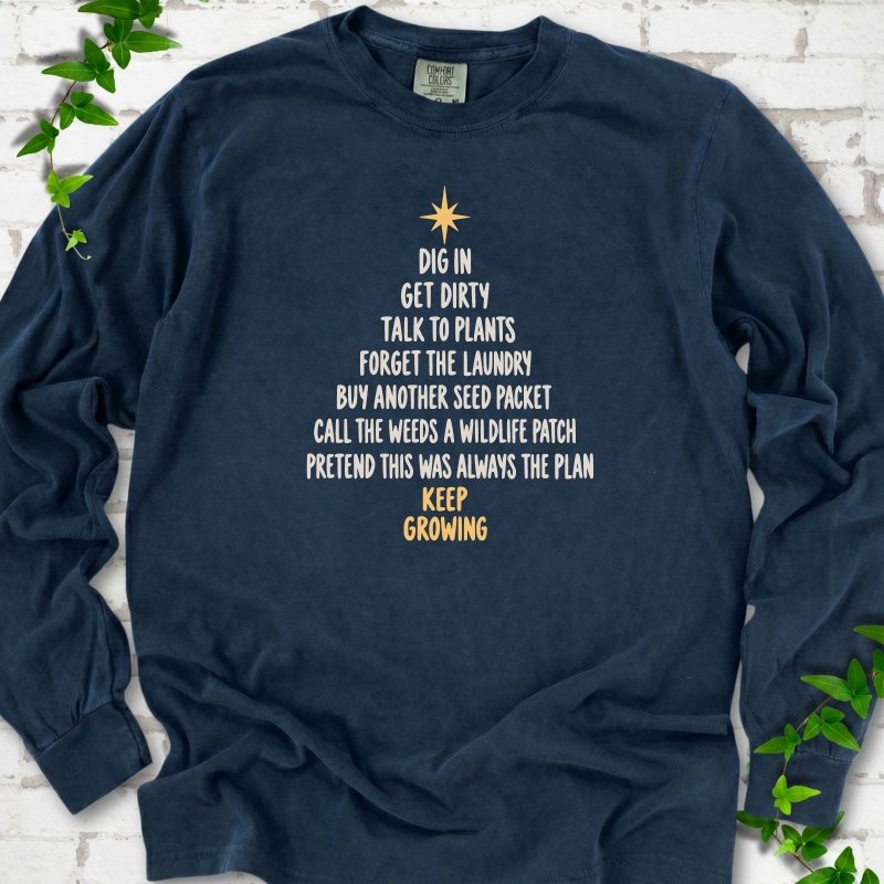 Midnight navy long sleeve T-shirt with a Christmas tree made of cream hand-lettered gardening phrases topped by a gold star, flat lay on white brick with ivy