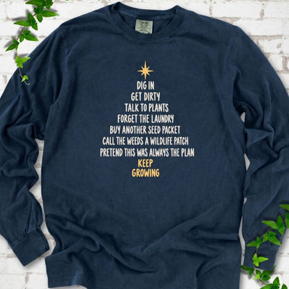 Midnight navy long sleeve T-shirt with a Christmas tree made of cream hand-lettered gardening phrases topped by a gold star, flat lay on white brick with ivy