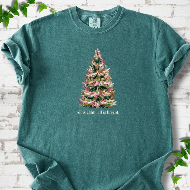 Blue Spruce T-shirt laid flat, featuring a decorated evergreen tree with pink ribbon garlands, a pink bow and gold ornaments above the words 'All is calm, all is bright.'
