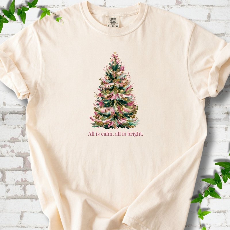 Ivory T-shirt laid flat, featuring a decorated evergreen tree with pink ribbon garlands, a pink bow and gold ornaments above the words 'All is calm, all is bright.'