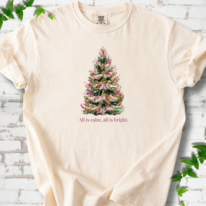 Ivory T-shirt laid flat, featuring a decorated evergreen tree with pink ribbon garlands, a pink bow and gold ornaments above the words 'All is calm, all is bright.'