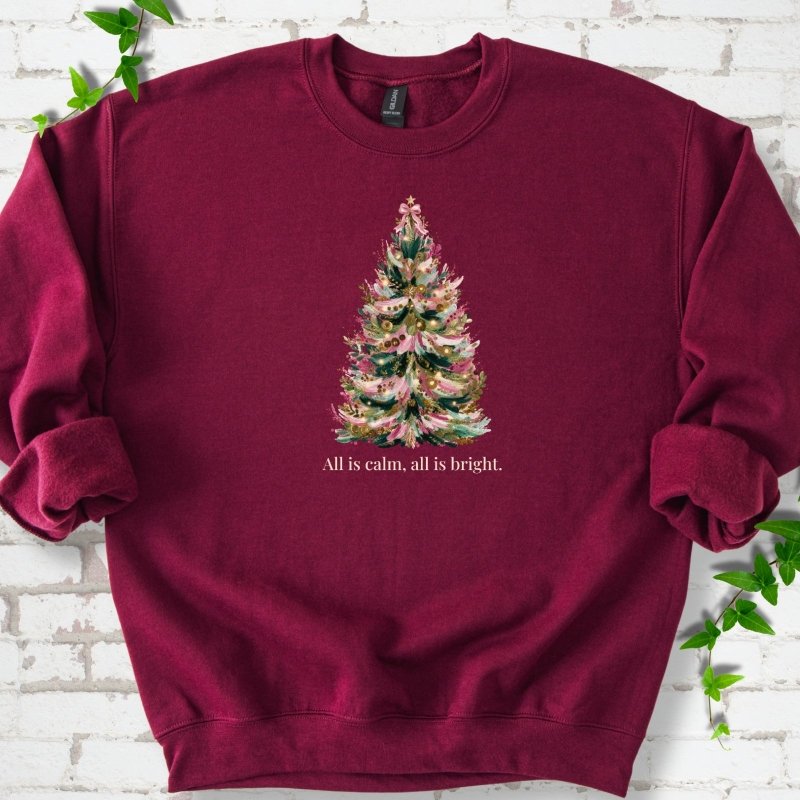 Maroon sweatshirt laid flat, featuring a decorated evergreen tree with pink ribbon garlands, a pink bow and gold ornaments above the words 'All is calm, all is bright.'