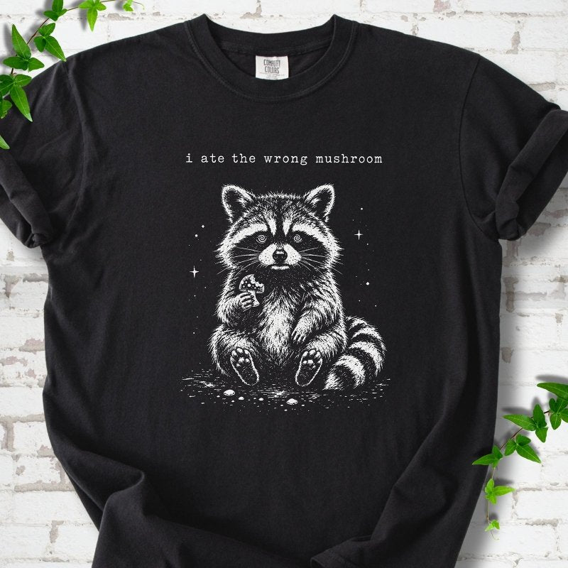 Black T-shirt flat lay with a white stippled illustration of a fluffy raccoon sitting up and holding a small mushroom, with small sparkles, below the lowercase typewriter-style words "i ate the wrong mushroom", on white brick with ivy sprigs.
