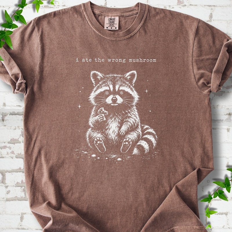 Espresso T-shirt flat lay with a white stippled illustration of a fluffy raccoon sitting up and holding a small mushroom, with small sparkles, below the lowercase typewriter-style words "i ate the wrong mushroom", on white brick with ivy sprigs.