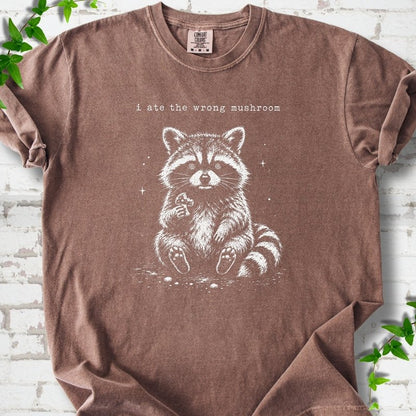 Espresso T-shirt flat lay with a white stippled illustration of a fluffy raccoon sitting up and holding a small mushroom, with small sparkles, below the lowercase typewriter-style words "i ate the wrong mushroom", on white brick with ivy sprigs.