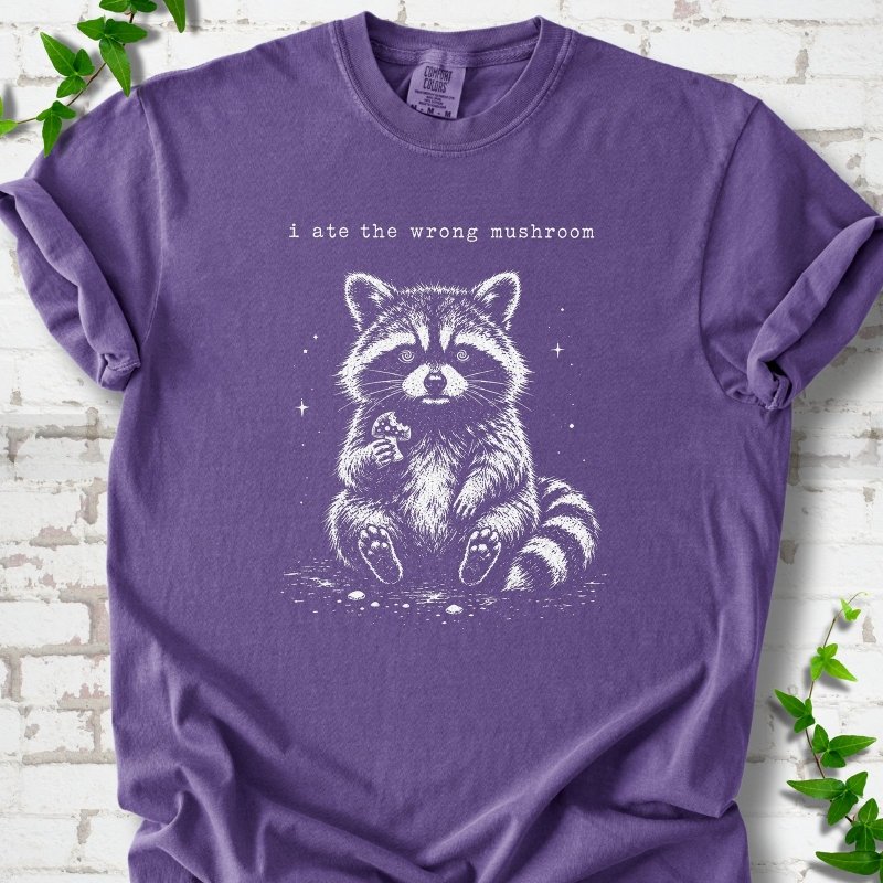 Grape T-shirt flat lay with a white stippled illustration of a fluffy raccoon sitting up and holding a small mushroom, with small sparkles, below the lowercase typewriter-style words "i ate the wrong mushroom", on white brick with ivy sprigs.