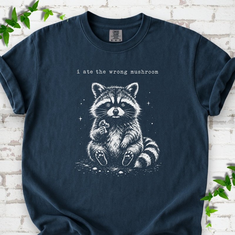 Midnight T-shirt flat lay with a white stippled illustration of a fluffy raccoon sitting up and holding a small mushroom, with small sparkles, below the lowercase typewriter-style words "i ate the wrong mushroom", on white brick with ivy sprigs.