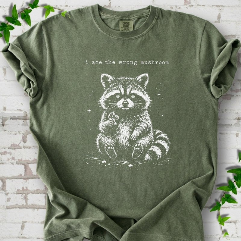 Moss T-shirt flat lay with a white stippled illustration of a fluffy raccoon sitting up and holding a small mushroom, with small sparkles, below the lowercase typewriter-style words "i ate the wrong mushroom", on white brick with ivy sprigs.