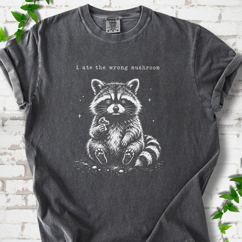 Pepper T-shirt flat lay with a white stippled illustration of a fluffy raccoon sitting up and holding a small mushroom, with small sparkles, below the lowercase typewriter-style words "i ate the wrong mushroom", on white brick with ivy sprigs.