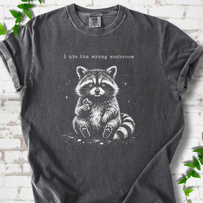 Pepper T-shirt flat lay with a white stippled illustration of a fluffy raccoon sitting up and holding a small mushroom, with small sparkles, below the lowercase typewriter-style words "i ate the wrong mushroom", on white brick with ivy sprigs.
