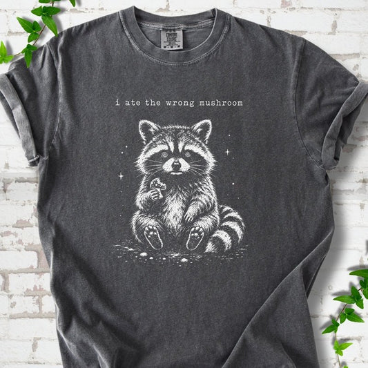 Pepper T-shirt flat lay with a white stippled illustration of a fluffy raccoon sitting up and holding a small mushroom, with small sparkles, below the lowercase typewriter-style words "i ate the wrong mushroom", on white brick with ivy sprigs.