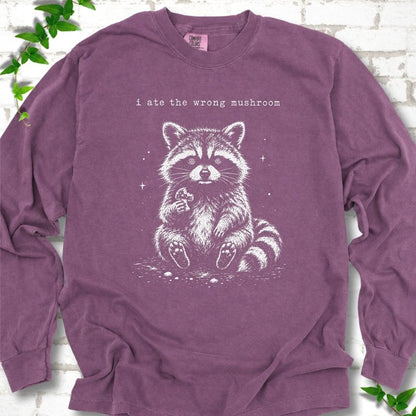 Berry long sleeve T-shirt flat lay with a white stippled illustration of a fluffy raccoon sitting up and holding a small mushroom, with small sparkles, below the lowercase typewriter-style words "i ate the wrong mushroom", on white brick with ivy sprigs.