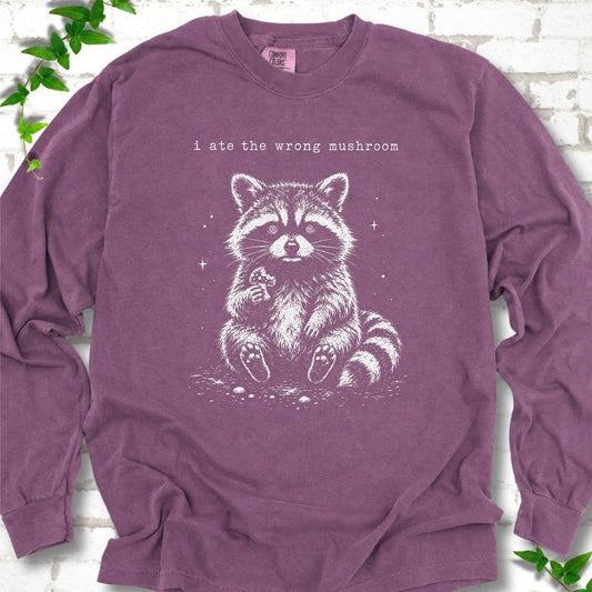 Berry long sleeve T-shirt flat lay with a white stippled illustration of a fluffy raccoon sitting up and holding a small mushroom, with small sparkles, below the lowercase typewriter-style words "i ate the wrong mushroom", on white brick with ivy sprigs.