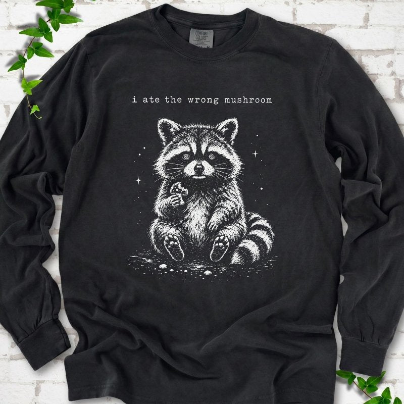 Black long sleeve T-shirt flat lay with a white stippled illustration of a fluffy raccoon sitting up and holding a small mushroom, with small sparkles, below the lowercase typewriter-style words "i ate the wrong mushroom", on white brick with ivy sprigs.