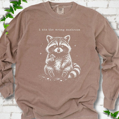 Espresso long sleeve T-shirt flat lay with a white stippled illustration of a fluffy raccoon sitting up and holding a small mushroom, with small sparkles, below the lowercase typewriter-style words "i ate the wrong mushroom", on white brick with ivy sprigs.
