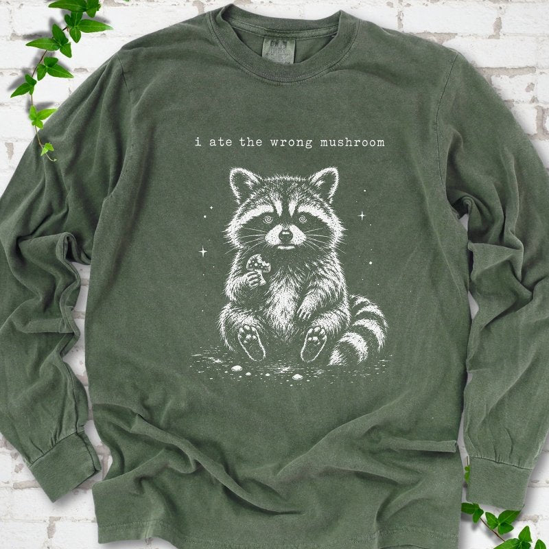 Hemp long sleeve T-shirt flat lay with a white stippled illustration of a fluffy raccoon sitting up and holding a small mushroom, with small sparkles, below the lowercase typewriter-style words "i ate the wrong mushroom", on white brick with ivy sprigs.