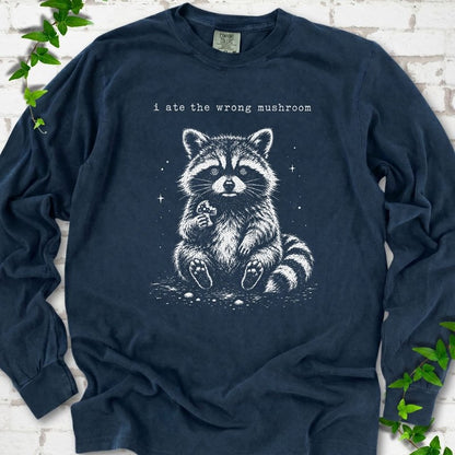 Midnight long sleeve T-shirt flat lay with a white stippled illustration of a fluffy raccoon sitting up and holding a small mushroom, with small sparkles, below the lowercase typewriter-style words "i ate the wrong mushroom", on white brick with ivy sprigs.