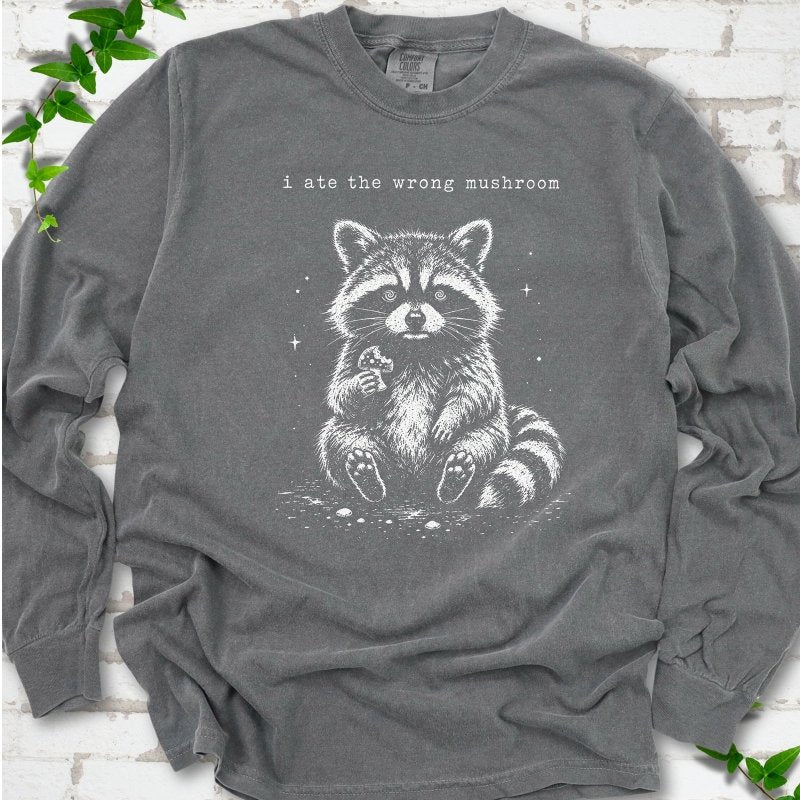 Pepper long sleeve T-shirt flat lay with a white stippled illustration of a fluffy raccoon sitting up and holding a small mushroom, with small sparkles, below the lowercase typewriter-style words "i ate the wrong mushroom", on white brick with ivy sprigs.