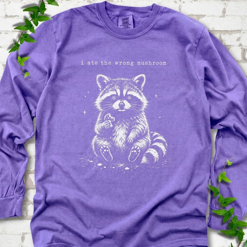 Violet long sleeve T-shirt flat lay with a white stippled illustration of a fluffy raccoon sitting up and holding a small mushroom, with small sparkles, below the lowercase typewriter-style words "i ate the wrong mushroom", on white brick with ivy sprigs.