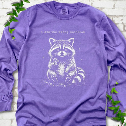 Violet long sleeve T-shirt flat lay with a white stippled illustration of a fluffy raccoon sitting up and holding a small mushroom, with small sparkles, below the lowercase typewriter-style words "i ate the wrong mushroom", on white brick with ivy sprigs.