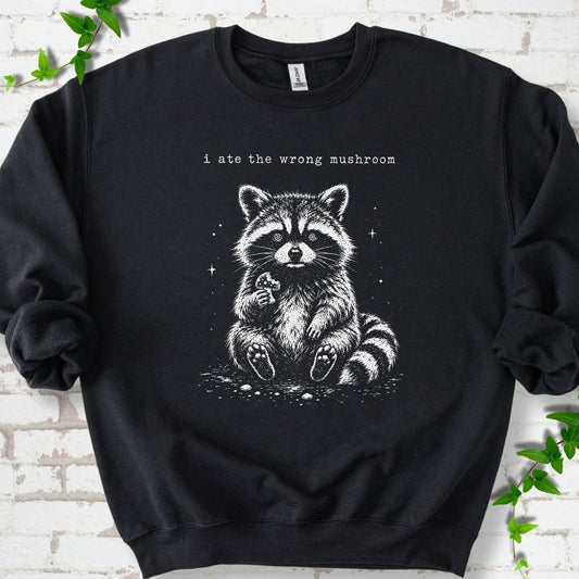 Black sweatshirt flat lay with a white stippled illustration of a fluffy raccoon sitting up and holding a small mushroom, with small sparkles, below the lowercase typewriter-style words "i ate the wrong mushroom", on white brick with ivy sprigs.