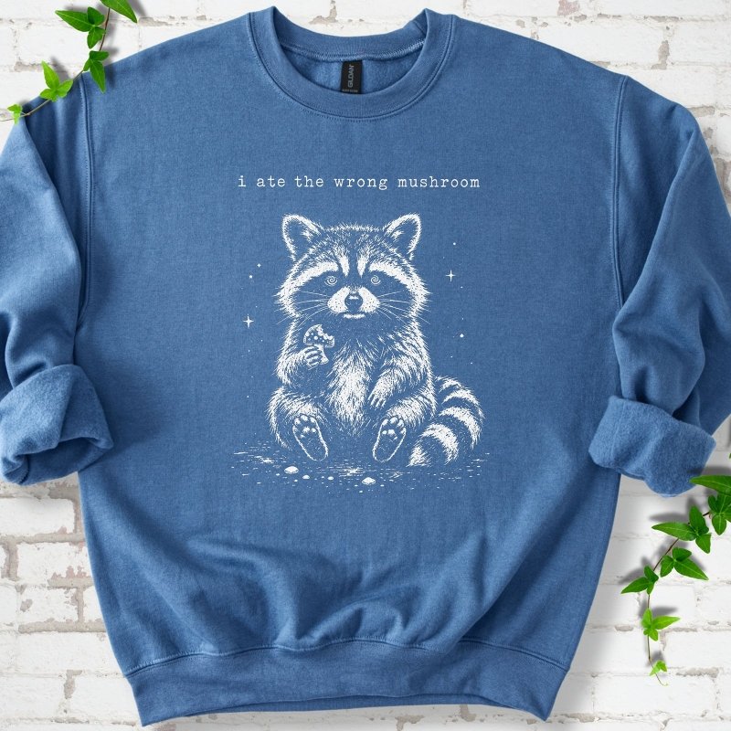 Indigo Blue sweatshirt flat lay with a white stippled illustration of a fluffy raccoon sitting up and holding a small mushroom, with small sparkles, below the lowercase typewriter-style words "i ate the wrong mushroom", on white brick with ivy sprigs.