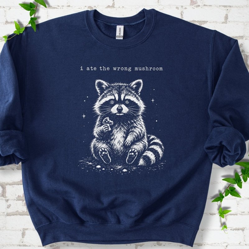 Navy sweatshirt flat lay with a white stippled illustration of a fluffy raccoon sitting up and holding a small mushroom, with small sparkles, below the lowercase typewriter-style words "i ate the wrong mushroom", on white brick with ivy sprigs.